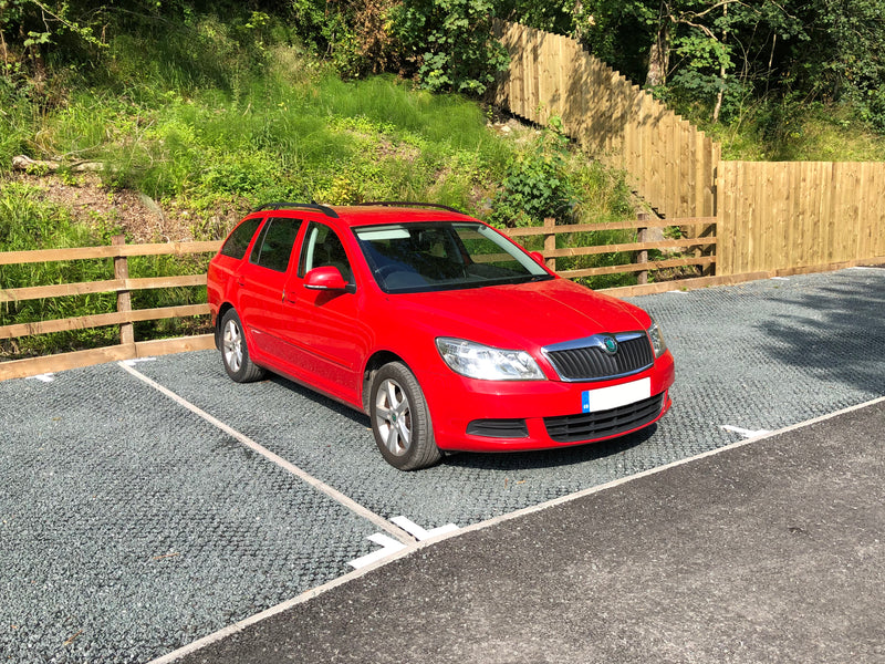 Load image into Gallery viewer, Red car parked on a surface of Bodpave 85 filled with gravel. A wooden fence and greenery in the background